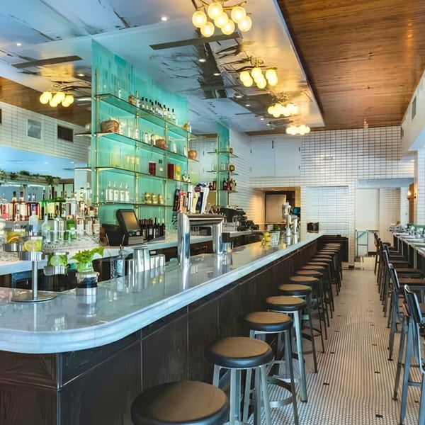 A long, empty bar with stools, a marble countertop, and shelves of bottles against a teal wall.