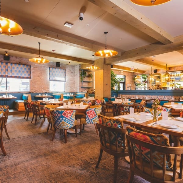 Spacious restaurant dining room with patterned chairs, wooden tables, brick walls, and a full bar.