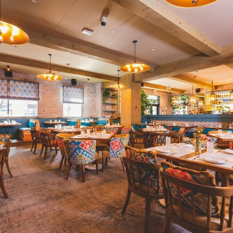 Spacious restaurant dining room with patterned chairs, wooden tables, brick walls, and a full bar.