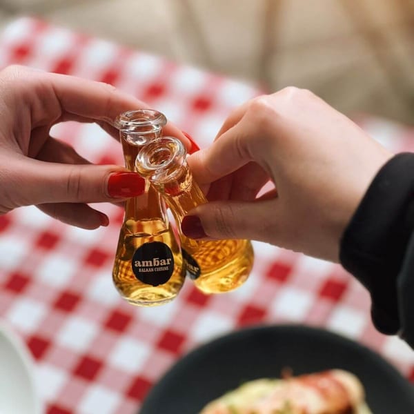 Two hands clinking small bottles of Ambar Balkan Cuisine liquor over a red and white checkered tablecloth.