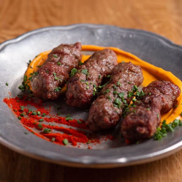 Four cevapi sausages on a pewter plate with orange sauce, red paprika, and chives.