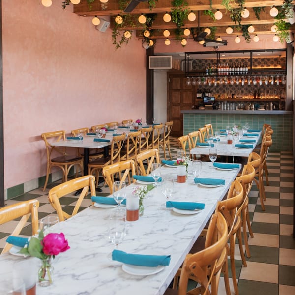 Long marble tables with blue napkins and wooden chairs in a restaurant with a bar and checkered floor.