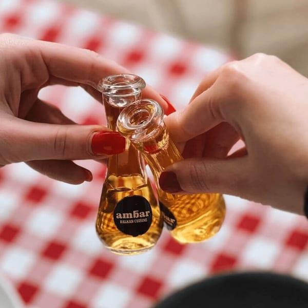Two hands clinking small bottles of Ambar Balkan Cuisine liquor over a red and white checkered tablecloth.