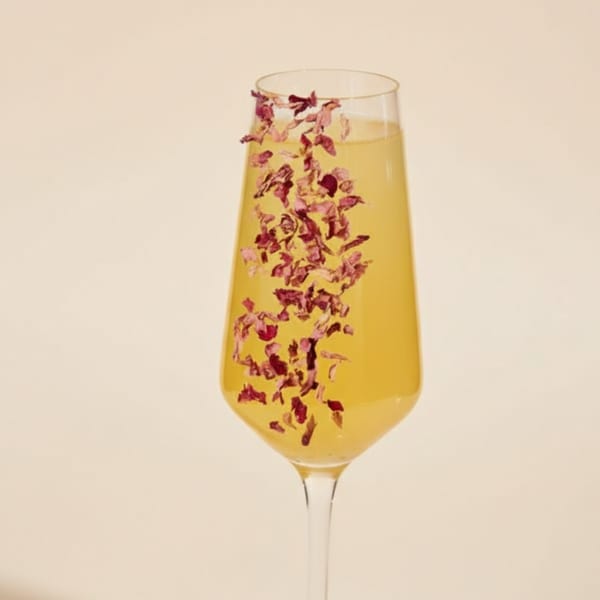 A champagne flute filled with a yellow drink and garnished with pink flower petals.