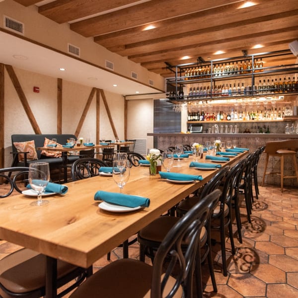 A long wooden dining table set with blue napkins and wine glasses, with a bar in the background.