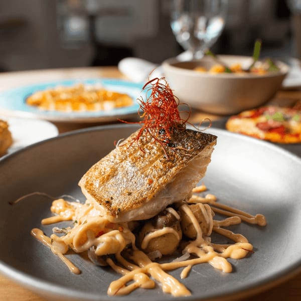 Crispy-skinned fish fillet with red garnish, creamy sauce, and potatoes on a gray plate at AMBAR.