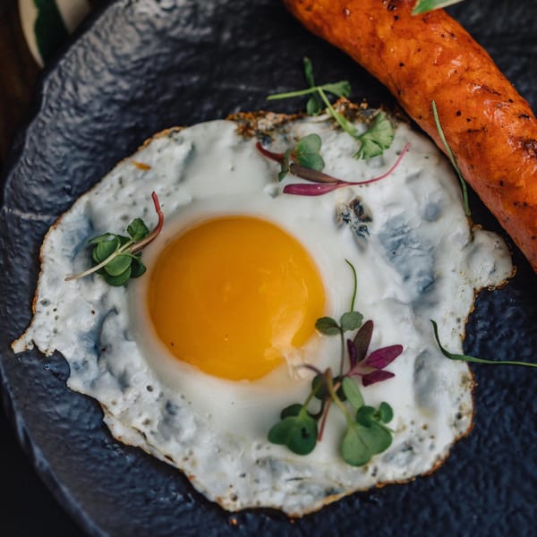 A close-up of a sunny-side-up egg with microgreens and a sausage on a dark plate.