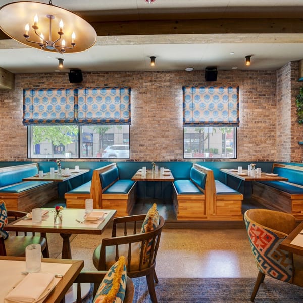 Restaurant interior with blue booths, wooden tables, brick walls, and patterned window shades.