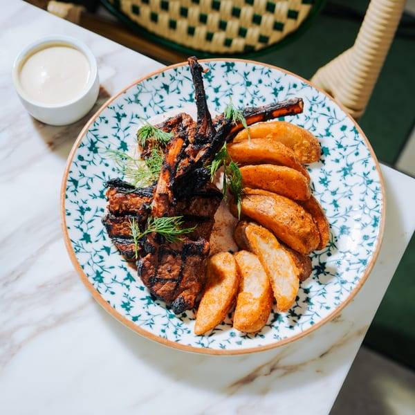 Grilled lamb chops with roasted potato wedges and fresh dill on a patterned plate.