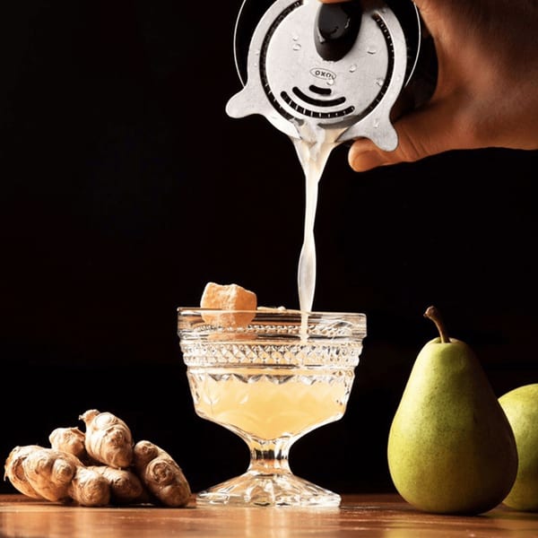 A bartender pours a light-colored cocktail into a decorative glass, with ginger and pears nearby.