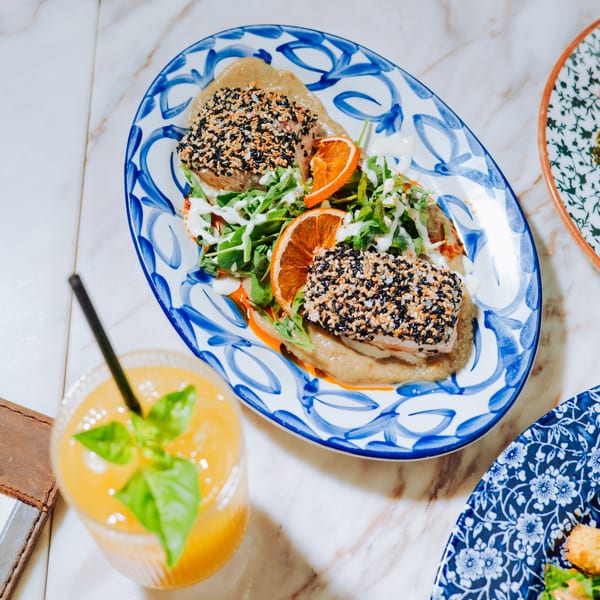 Two sesame-crusted fish fillets with arugula and orange on a blue patterned plate, beside an orange drink.