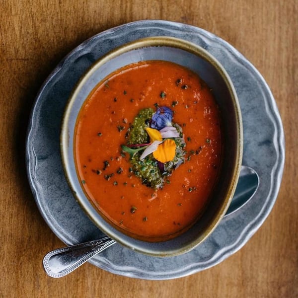Overhead view of a bowl of red tomato soup with green pesto and edible flowers.