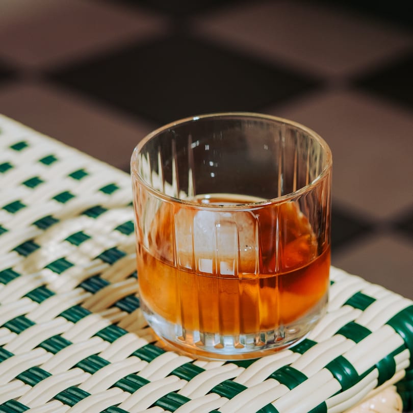 Amber drink with a large ice cube in a ribbed glass on a white and green woven surface.