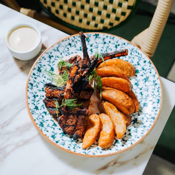 Grilled lamb chops and potato wedges on a patterned plate with a side of dipping sauce.