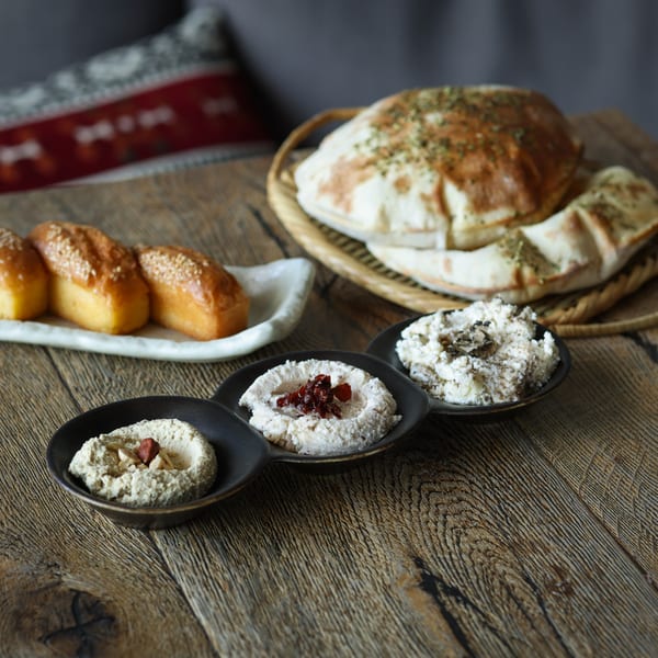 Three small bowls of dips, pita bread in a basket, and sesame bread on a wooden table.