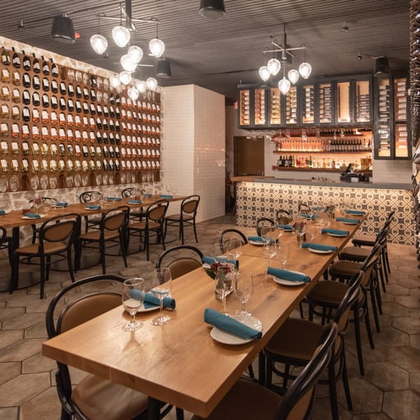 Restaurant dining room with long wooden tables, wine bottle displays, and a bar with patterned tiles.