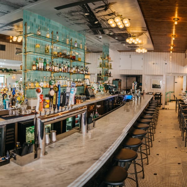 A long, empty bar with a marble countertop, bar stools, and shelves of liquor bottles.