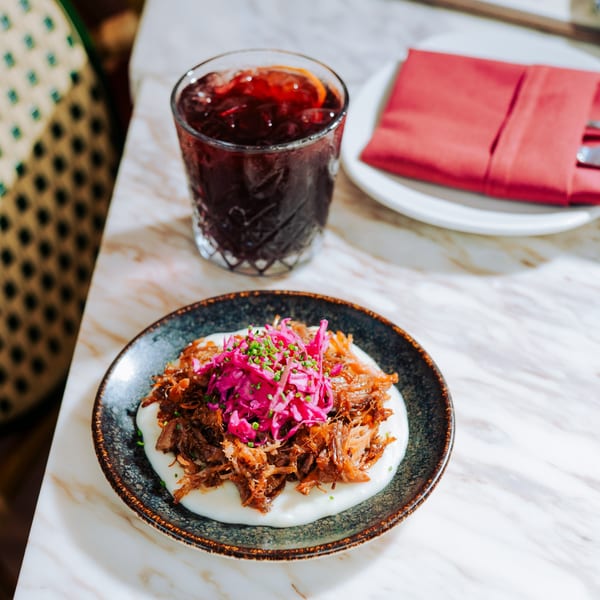 Pulled pork with pickled red cabbage and chives on a white sauce, next to a dark red drink.