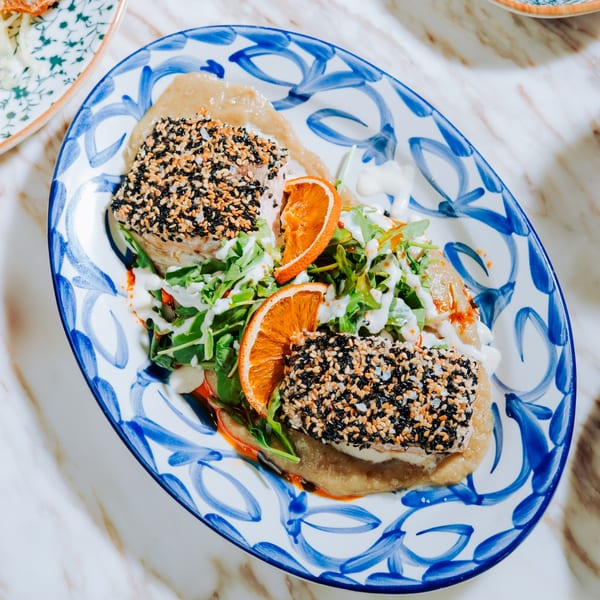 Two sesame-crusted fish fillets with arugula, orange slices, and sauce on a blue and white plate.