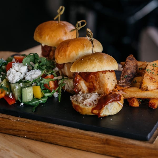 Three sliders with barbecue sauce, coleslaw, and fries, served with a side salad on a dark plate.