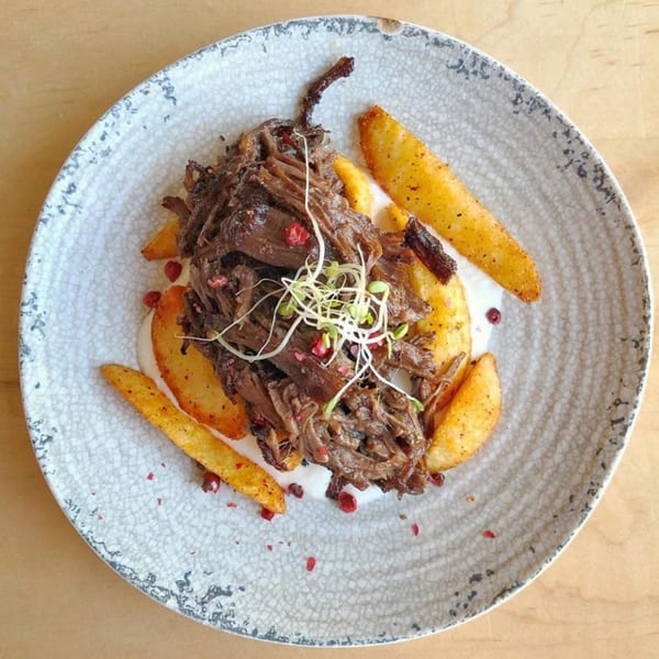 Shredded European-style beef with potato wedges, white sauce, and microgreens on a textured plate.