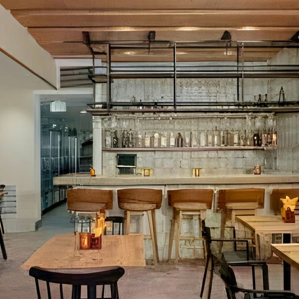 Industrial-chic restaurant interior with a concrete bar, wooden stools, and exposed brick, featuring bottles and glasses.