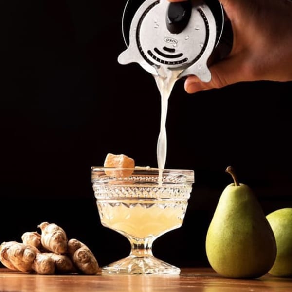 A hand pours a light-colored cocktail from a shaker into a crystal glass with ginger and pears.