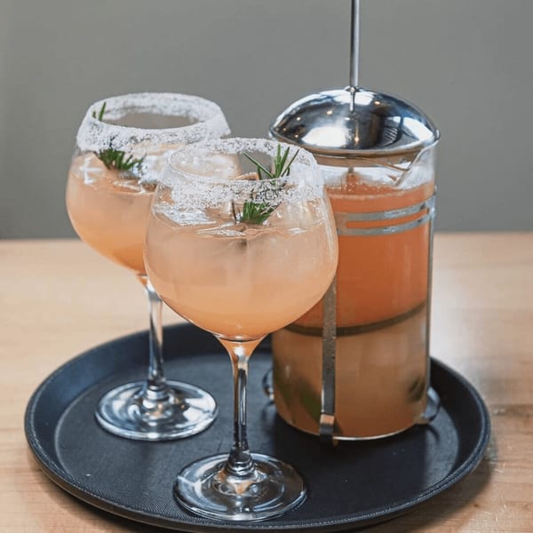 Two sugar-rimmed cocktails with rosemary and a French press of European drinks on a black tray.