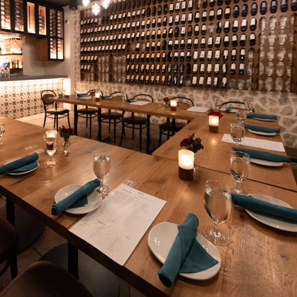 Restaurant dining room with wooden tables, teal napkins, candles, and a wall of wine bottles.